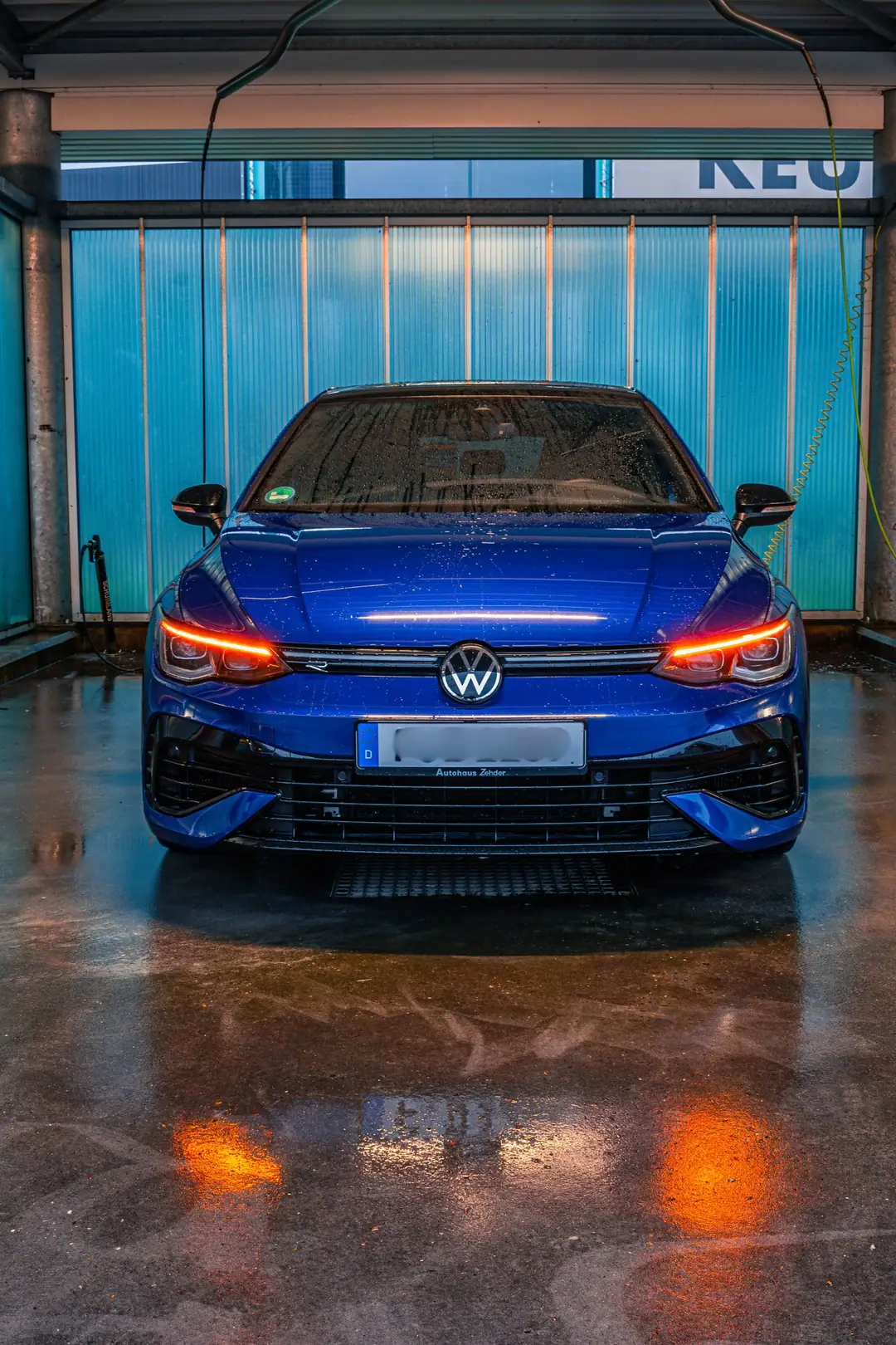 Blauwe Volkswagen Golf R recht van voren in een wasbox met brandende ledverlichting en oranje reflecties op de natte vloer
