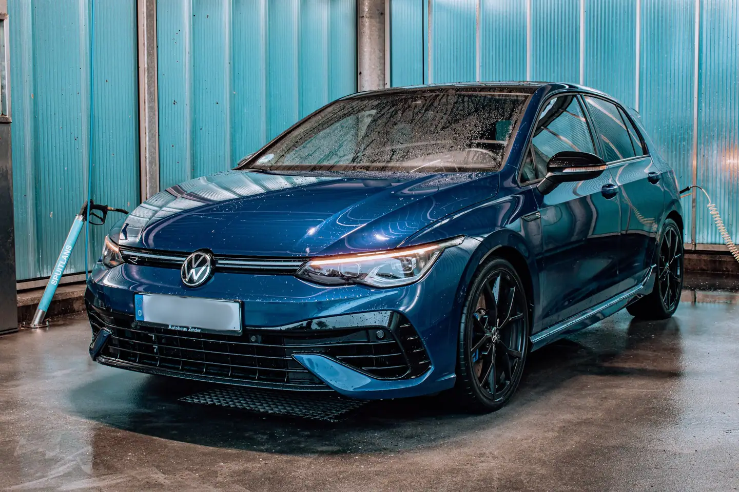 Natte blauwe Volkswagen Golf R schuin van voren in een wasbox met turquoise wanden en een hogedrukspuit