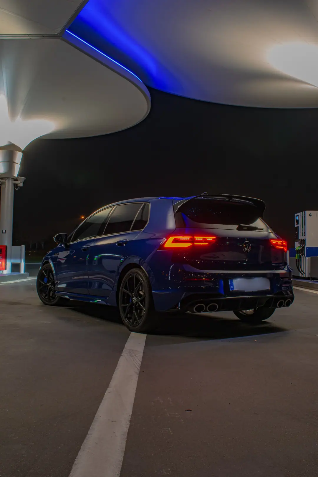 Blauwe Volkswagen Golf R schuin van achteren bij nacht onder een verlichte overkapping met blauwe ledrand