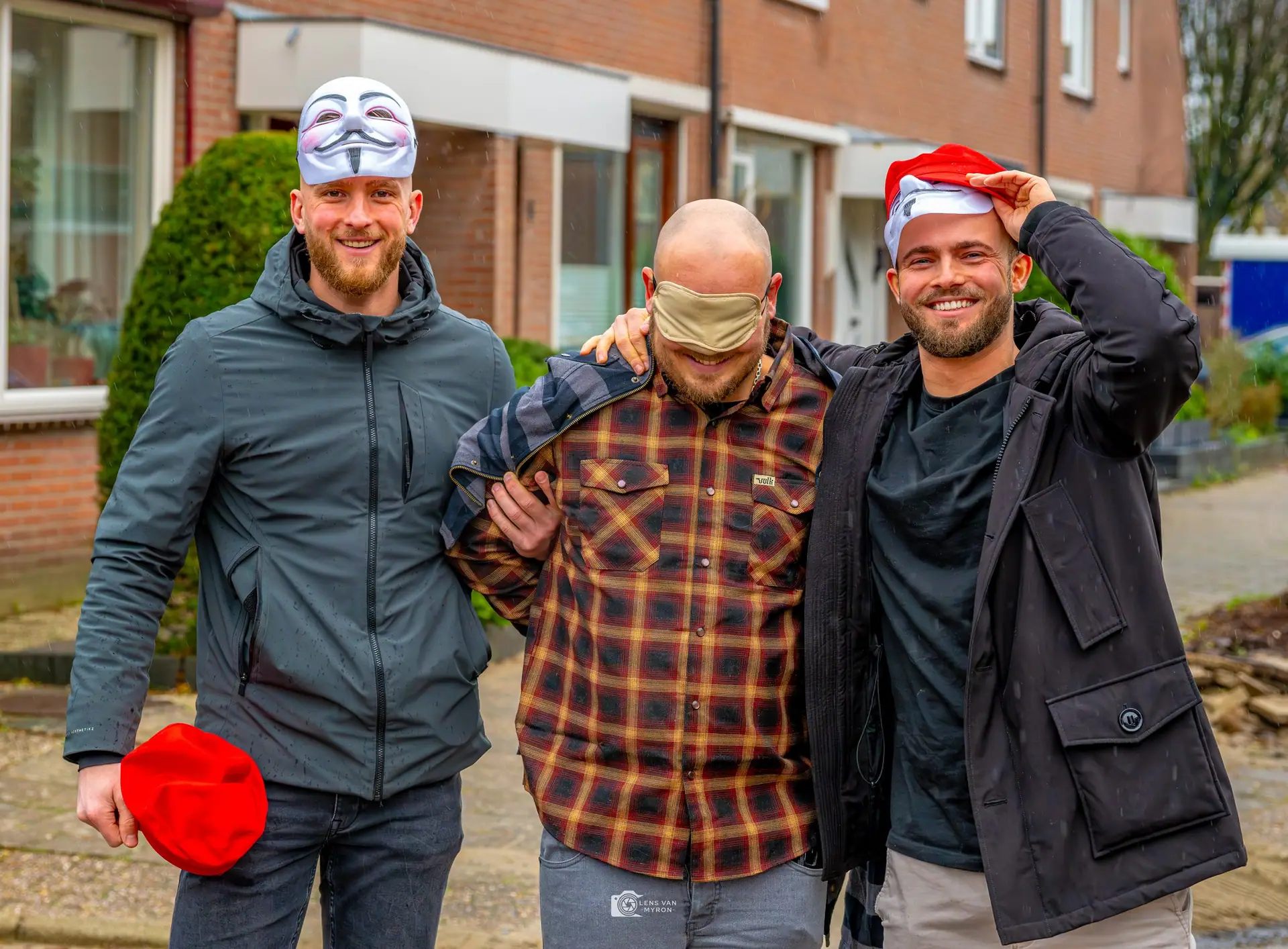 Drie vrienden in een woonstraat; de vrijgezel in het midden is geblinddoekt, links een vriend met een masker op zijn hoofd, rechts een vriend met een kerstmuts.