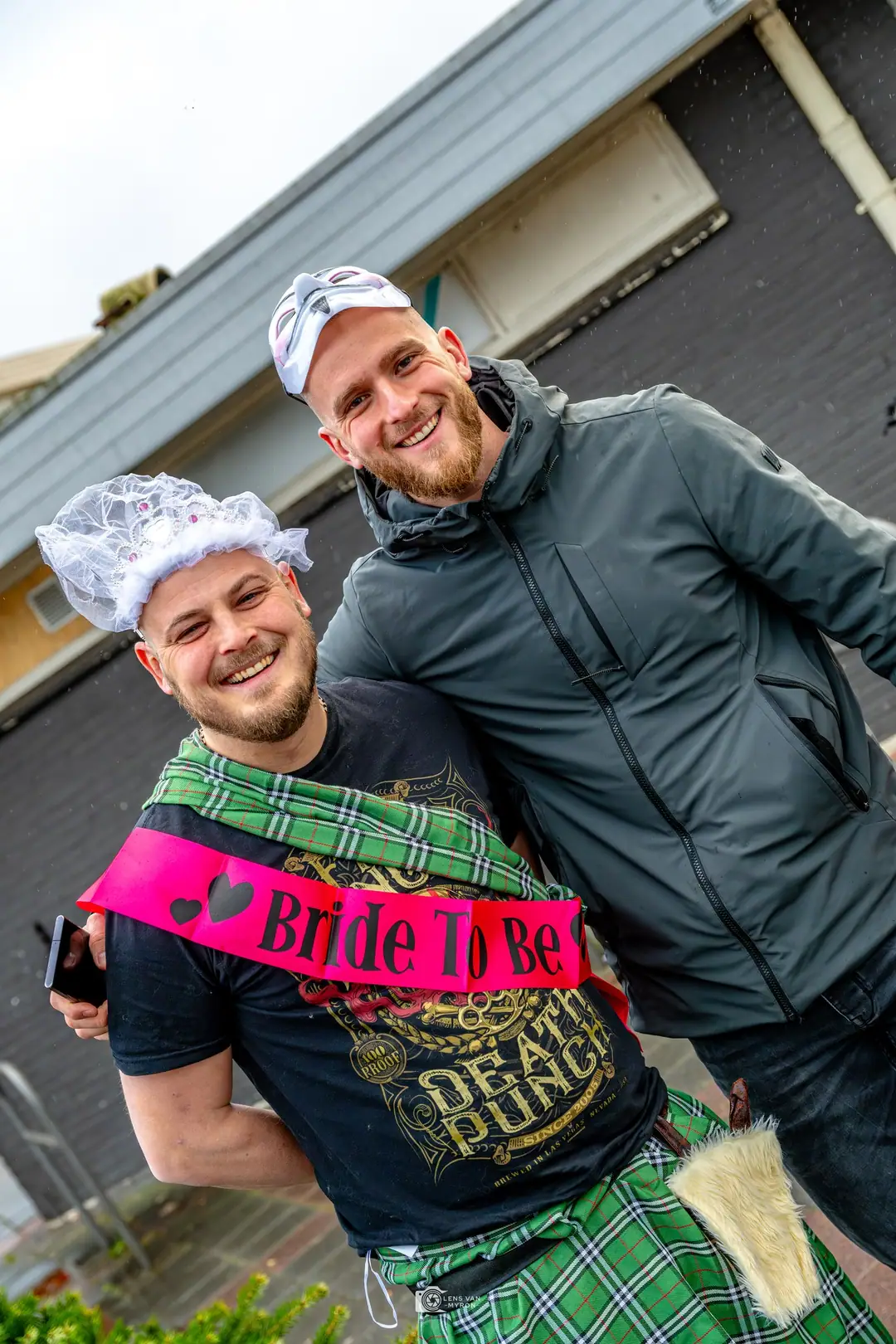 De vrijgezel lacht in een kilt, met een sluiertje op en een roze "Bride To Be"-sjerp; een vriend slaat een arm om hem heen.