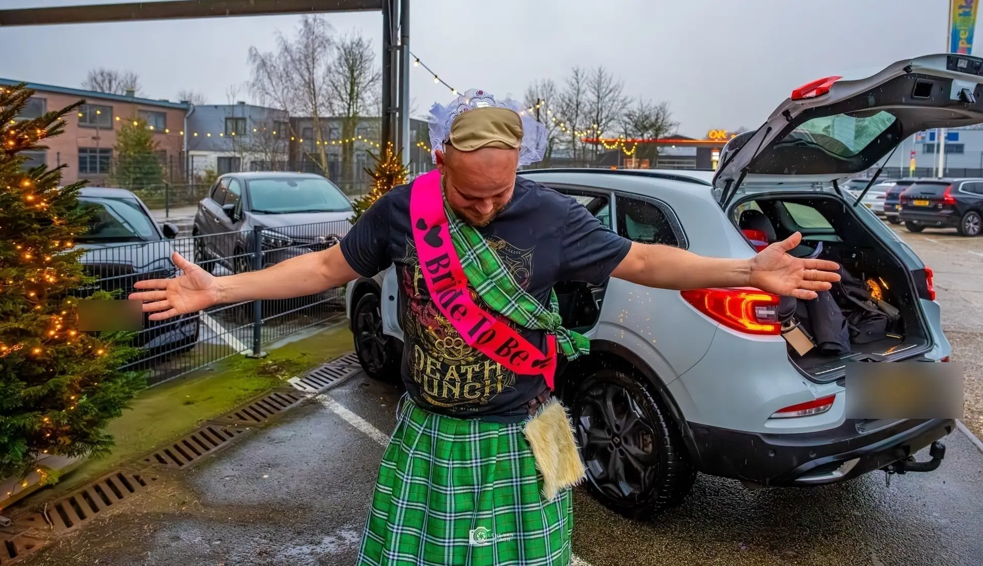 De vrijgezel in kilt en roze sjerp spreidt zijn armen op een parkeerplaats met kerstlichtjes en een versierde kerstboom.