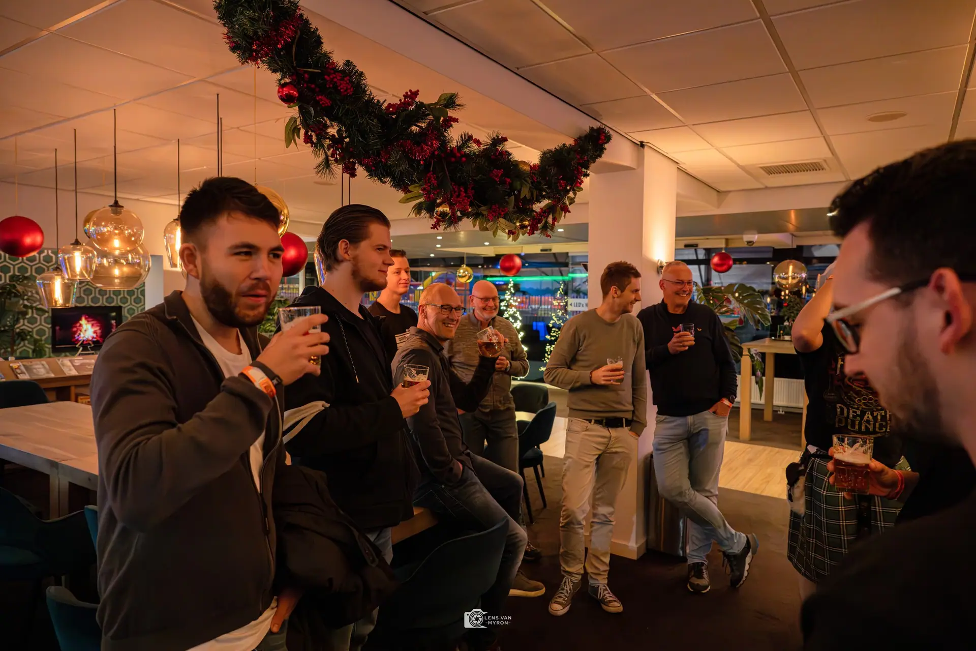 De groep staat met een biertje aan de borrel onder een kerstslinger met rode ballen.